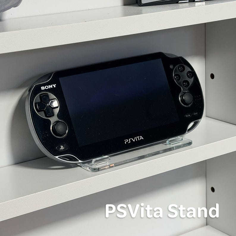 May include: Black PS Vita handheld console on a clear acrylic stand, placed on a white shelf. The console features the Sony logo and "PSVita" text. The stand is labelled "PSVita Stand".