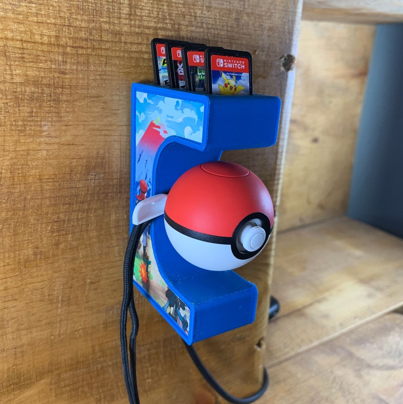 May include: A blue and white 3D printed wall mount shaped like the number 5 with a Pokeball holder and a slot for Nintendo Switch game cartridges. The wall mount has a black lanyard attached to it.