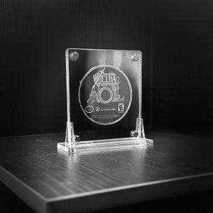 May include: A clear acrylic display case holding a Zelda GameCube disc. The disc is visible through the transparent case, which is secured with four small screws. The case is on a dark wooden surface.