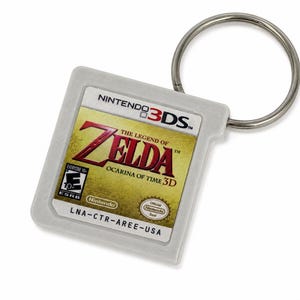 May include: A square, light grey plastic keyring shaped like a Nintendo 3DS game cartridge. The label features the gold-toned title The Legend of Zelda Ocarina of Time 3D, the Nintendo 3DS logo, an ESRB Everyone 10+ rating, and the code LNA-CTR-AREE-USA.