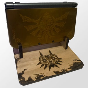 May include: A gold-colored handheld gaming console with a Triforce design rests on a wooden stand. The stand features a Majora's Mask design and other etched details. The console is black and gold.