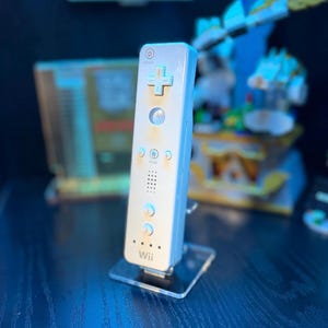 Puede incluir: Un mando a distancia Wii blanco con una cruceta direccional en forma de cruz y varios botones. El mando está montado en un soporte de acrílico transparente. La palabra "Wii" está impresa en la parte inferior. El fondo está borroso, mostrando otros objetos.