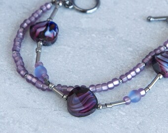 May include: A double-strand bracelet with silver-toned metal accents and purple and blue glass beads. The bracelet features three larger, heart-shaped beads with a swirling purple and white pattern.