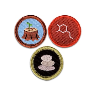 May include: Three circular embroidered patches. The top left patch has a light blue background with a brown tree stump and a small green sprout. The top right patch is red with a white chemical structure. The bottom patch is black with three stacked light gray stones and a gold border.