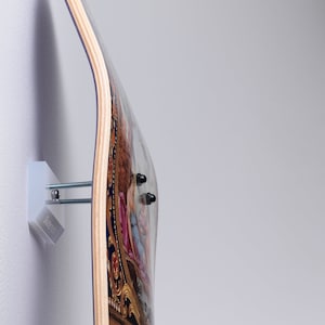 Op de afbeelding: Een skateboard met een klassiek schilderijontwerp, gemonteerd aan een witte muur met een metalen beugel. Het skateboard heeft een houten kern en een gebogen vorm. De beugel heeft de tekst "SkateUp".