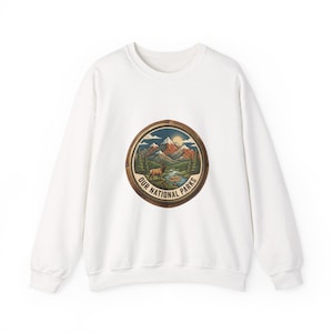 Op de afbeelding: Witte sweatshirt met een cirkelvormige afbeelding van een berglandschap, een hert en de tekst "OUR NATIONAL PARKS PROTECT". Het ontwerp doet denken aan vintage reisposters, met de nadruk op natuur en behoud.