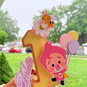 May include: A yellow and pink number one cake topper with a cartoon character holding balloons. The topper also features a bee on a flower and the name "Arlette" in cursive. Perfect for a first birthday celebration.