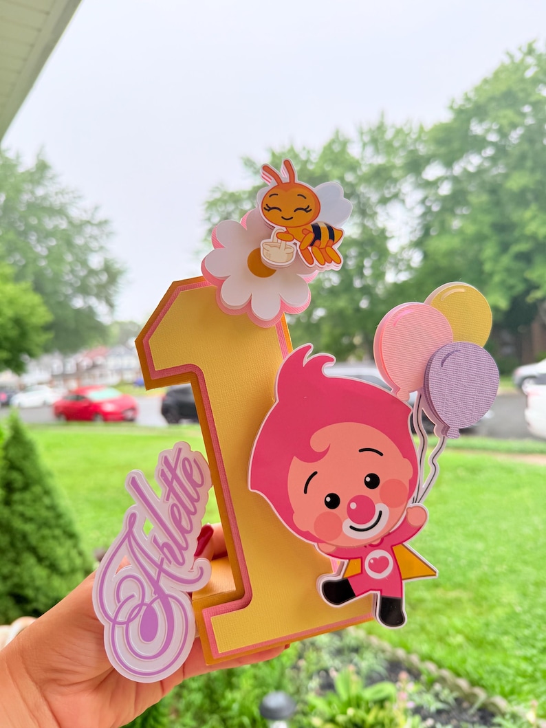 May include: A yellow and pink number one cake topper with a cartoon character holding balloons. The topper also features a bee on a flower and the name "Arlette" in cursive. Perfect for a first birthday celebration.