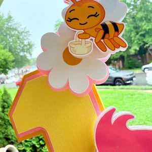 May include: A colorful birthday cake topper featuring a cartoon bee carrying a basket, perched on a daisy. The number "1" is in yellow with pink trim. The topper is designed for a first birthday celebration.
