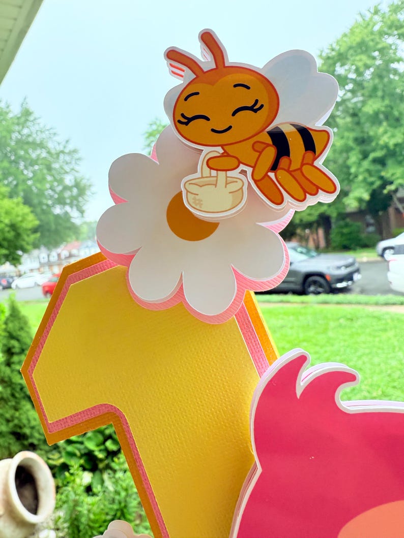 May include: A colorful birthday cake topper featuring a cartoon bee carrying a basket, perched on a daisy. The number "1" is in yellow with pink trim. The topper is designed for a first birthday celebration.