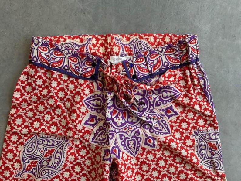 May include: Red and white patterned trousers with a purple and white floral design. The trousers have a drawstring waist and a decorative bow. The fabric is a soft, lightweight material, ideal for warmer weather.