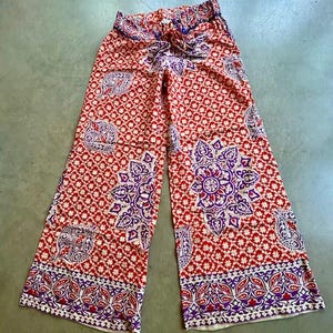 May include: Wide-legged palazzo trousers in a red and white pattern with floral and paisley motifs. The design incorporates purple and white details. The trousers have a drawstring waist.