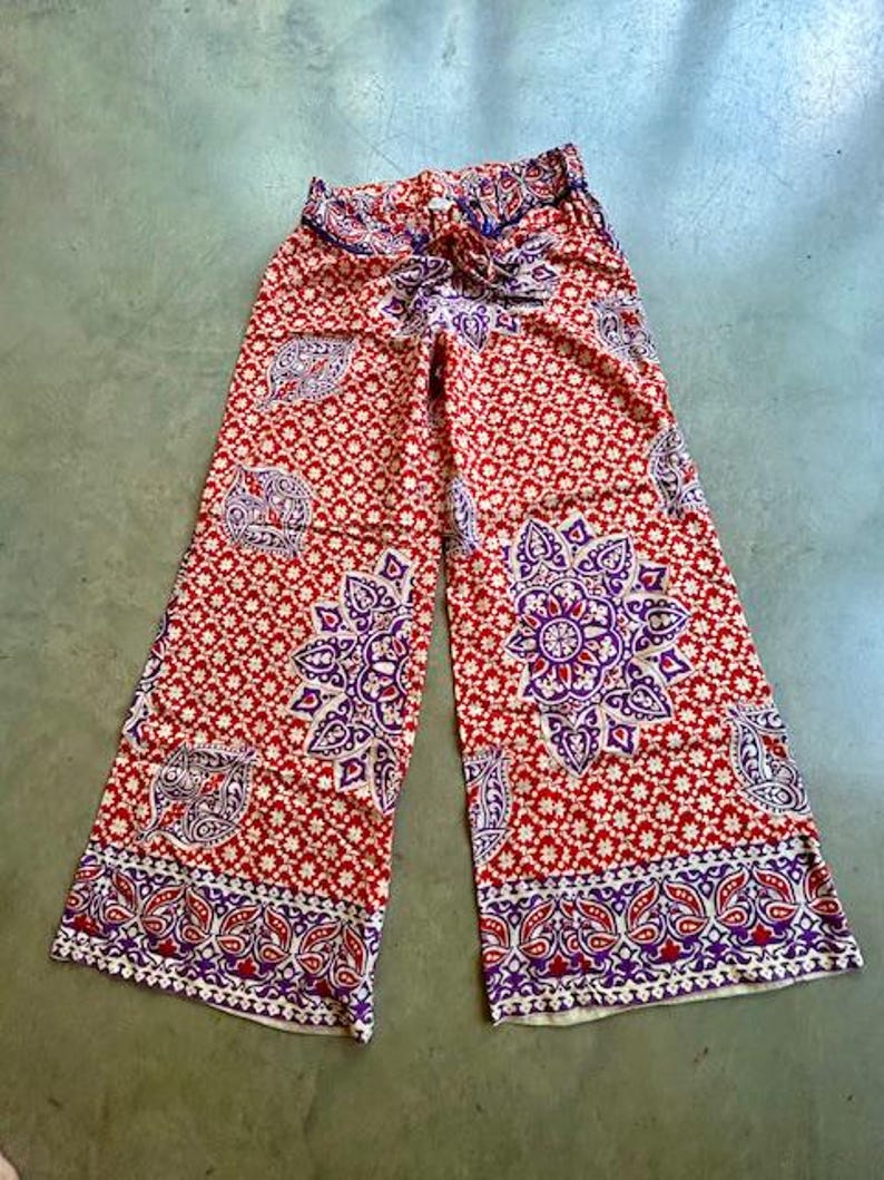 May include: Wide-legged palazzo trousers in a red and white pattern with floral and paisley motifs. The design incorporates purple and white details. The trousers have a drawstring waist.