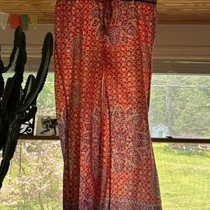 May include: Red and cream palazzo trousers with a floral and paisley pattern. The trousers are hanging from a hanger in front of a window. The fabric has a detailed design with a border at the bottom.