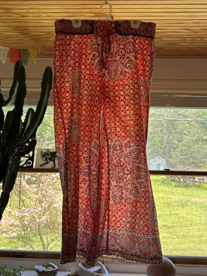 May include: Red and cream palazzo trousers with a floral and paisley pattern. The trousers are hanging from a hanger in front of a window. The fabric has a detailed design with a border at the bottom.
