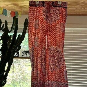 May include: Red and purple patterned palazzo trousers hanging on a white hanger. The trousers feature a floral and paisley design. The background includes a window, cactus, and colorful flags.