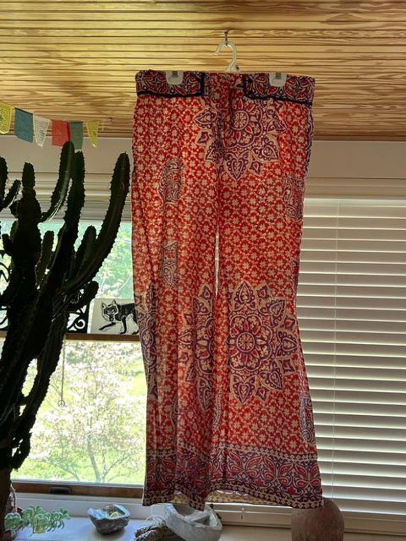 May include: Red and purple patterned palazzo trousers hanging on a white hanger. The trousers feature a floral and paisley design. The background includes a window, cactus, and colorful flags.