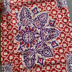 May include: Close-up of a red fabric with a white floral pattern and a large purple and white floral design in the centre. The fabric also features purple paisley designs in the corners.