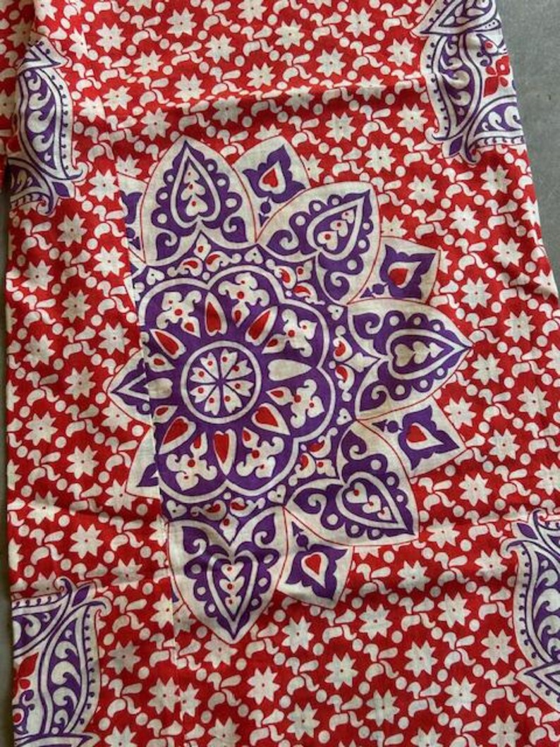 May include: Close-up of a red fabric with a white floral pattern and a large purple and white floral design in the centre. The fabric also features purple paisley designs in the corners.
