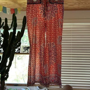 May include: A pair of red and cream patterned trousers hanging from a white hanger. The trousers display a repeating floral and paisley design. The fabric is lightweight, suitable for warm weather. The trousers are hanging in front of a window.