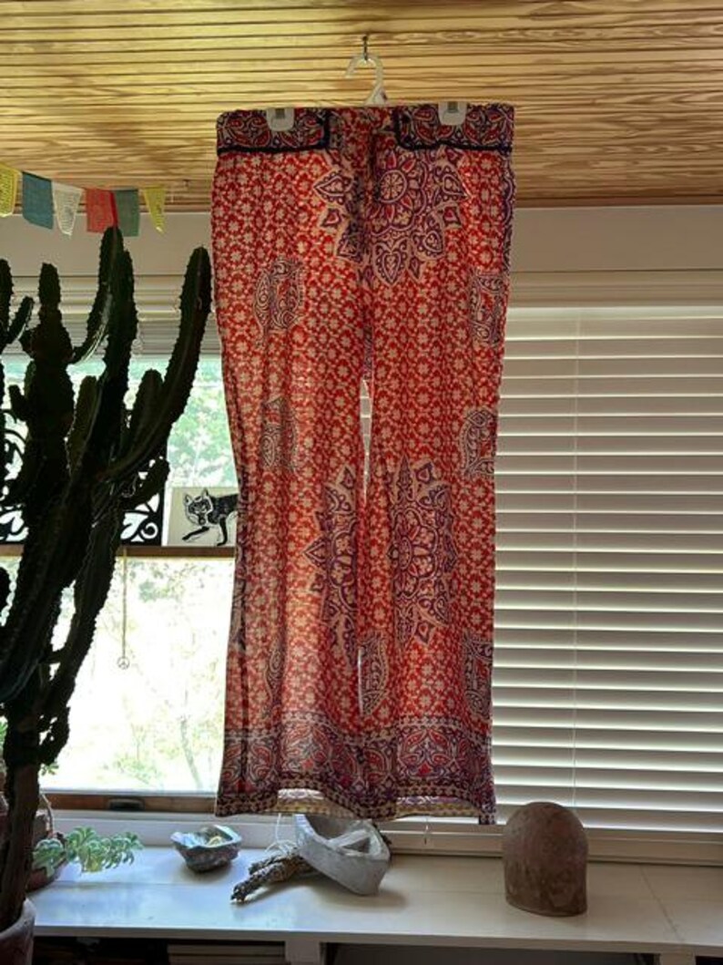 May include: A pair of red and cream patterned trousers hanging from a white hanger. The trousers display a repeating floral and paisley design. The fabric is lightweight, suitable for warm weather. The trousers are hanging in front of a window.