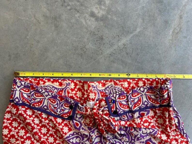 May include: Close-up of red and white patterned trousers with purple and blue trim. A yellow measuring tape is laid across the top, showing a measurement of approximately 33 cm.