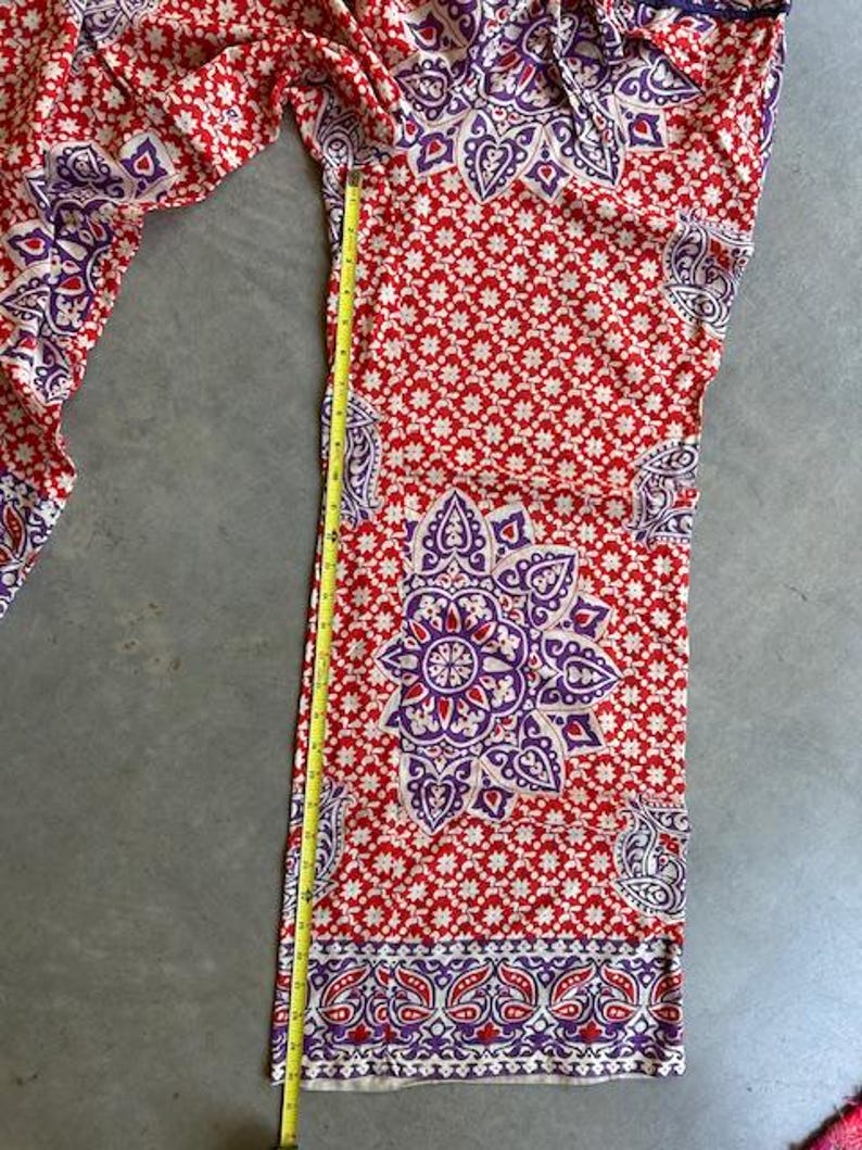 May include: Red and white patterned trousers with a purple floral design. The trousers are measured with a yellow tape measure. The trousers have a wide leg and a repeating pattern of small white flowers.
