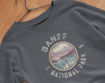 May include: A dark gray sweatshirt with a vintage-style graphic of a mountain scene and the text "Banff National Park Est. 1885".