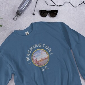 May include: A blue sweatshirt with a vintage-style graphic of Washington D.C. The graphic features a circular design with the text "WASHINGTON EST 1790 D.C." and a picture of a cityscape with cherry blossoms.