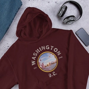 May include: Burgundy hooded sweatshirt with a graphic print of the Washington D.C. skyline with cherry blossoms. The text "WASHINGTON EST 1790 D.C." is printed on the sweatshirt.