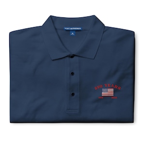 A folded navy blue Port Authority polo shirt with vibrant red embroidery on the left chest. The design features "250 YEARS" arched in red thread above an American flag, with the years "1776 – 2026" stitched in red below. The shirt has a structured collar and three dark buttons.