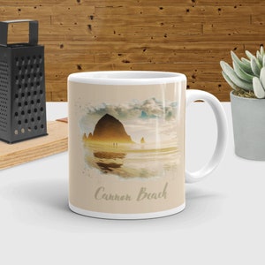 May include: White ceramic mug with a watercolor image of Cannon Beach, Oregon. The image features Haystack Rock, a large sea stack, and the Pacific Ocean. The text "Cannon Beach" is printed on the mug.