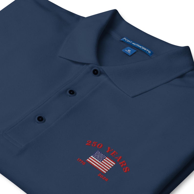 An angled close-up of a navy blue Port Authority polo shirt, showcasing the intricate embroidery on the chest. The design features a vibrant red arched "250 YEARS" and "1776 - 2026" surrounding a detailed American flag. The shot highlights the professional stitch work and the textured pique knit of the fabric.