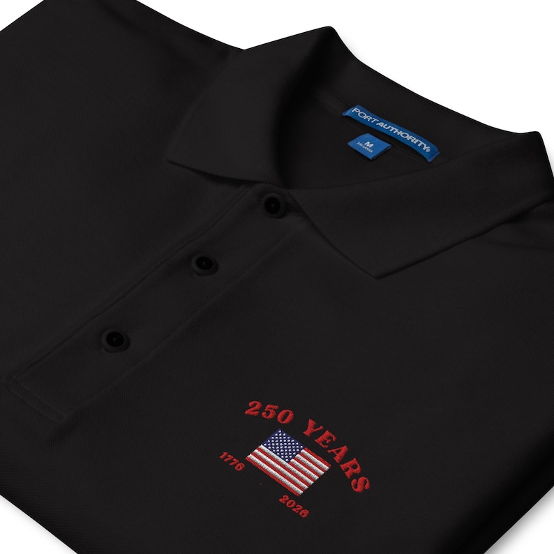 Close-up of embroidery on a black patriotic polo shirt for the USA 250th anniversary. Detailed stitching of the American flag and the years 1776 to 2026 in vibrant red thread. High-quality commemorative apparel for the American Semi quincentennial.