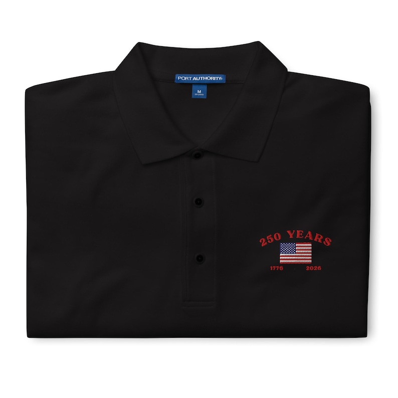 A folded black Port Authority polo shirt with sharp red embroidery on the left chest. The design features a bold red arched "250 YEARS" above a classic red, white, and blue American flag, with red "1776 – 2026" dates stitched below. The shirt includes a structured collar and three black buttons.