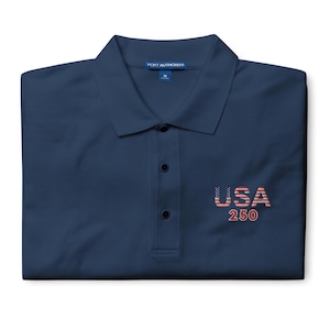 May include: Navy blue polo shirt with a collared neckline and three-button placket. The shirt features the text "USA 250" embroidered in red, white, and blue on the chest. The shirt is from the brand "Port Authority".