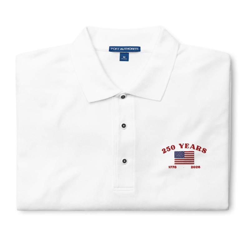 White embroidered polo shirt celebrating the USA 250th anniversary. The left-chest design showcases the American flag and the historic dates 1776-2026 in vibrant red thread. High-quality Port Authority brand men’s golf shirt for the 2026 Semi quincentennial.