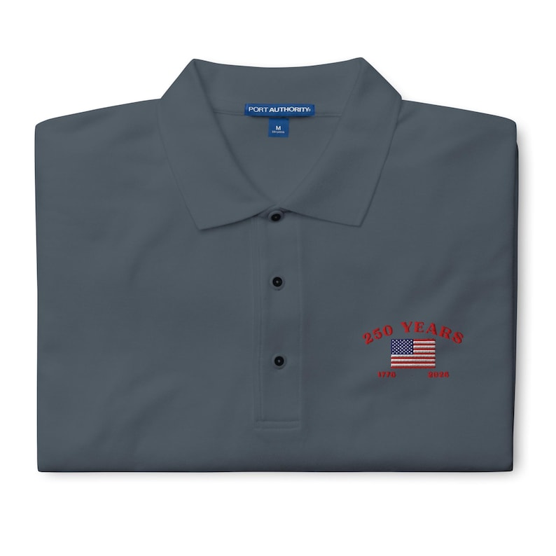 A folded charcoal grey Port Authority polo shirt featuring vibrant red embroidery on the left chest. The design shows a red arched "250 YEARS" above a classic American flag, with red "1776 - 2026" dates stitched below. The shirt has a structured collar and three dark buttons.
