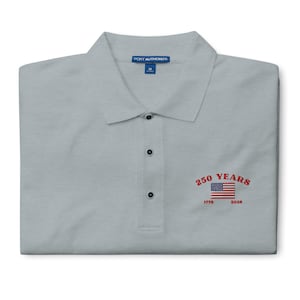 Close-up of a light grey polo shirt with a red embroidered 250th American anniversary logo, featuring the U.S. flag and the years 1776 to 2026.