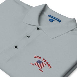 Angled close-up of a light grey polo shirt featuring red embroidered text "250 YEARS" and "1776 - 2026" wrapped around a classic American flag logo.
