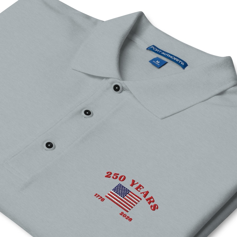Angled close-up of a light grey polo shirt featuring red embroidered text "250 YEARS" and "1776 - 2026" wrapped around a classic American flag logo.