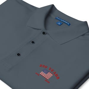 Close-up of red embroidery on a charcoal grey patriotic polo shirt for the USA 250th anniversary. Detailed stitching of the American flag and the years 1776 to 2026 in vibrant red thread. High-quality commemorative apparel for the American Semi quincentennial celebration.