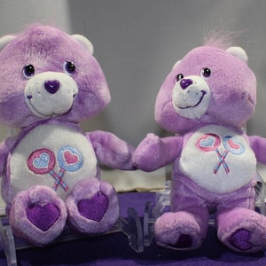 May include: Two purple Care Bears plush toys. The Care Bears are sitting on a purple surface. Both Care Bears have white bellies with pink and blue hearts on lollipops. The Care Bears have purple noses and black eyes.