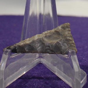 May include: A grey arrowhead with a rough, textured surface. The arrowhead is displayed on a clear plastic stand.