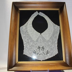 May include: A white lace collar framed in a dark wood frame. The lace is delicate and intricate, with a floral pattern. The collar is displayed on a black background.