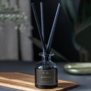 Puede incluir: Un difusor de varillas de aromaterapia de vidrio negro con varillas negras. La etiqueta dice "AVENTOS" con texto adicional. El difusor está sobre una tabla de madera, con un fondo oscuro.