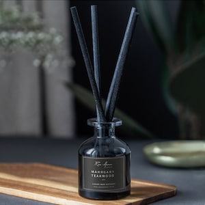 May include: A black glass reed diffuser with black reeds, labeled "Mahogany Teakwood." The diffuser sits on a wooden board. The label has the brand name "Rose Avenue" and the words "Luxury Reed Diffuser."