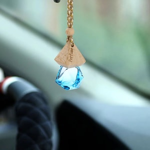 Op de afbeelding: Een hangende auto-luchtverfrisser met een gefacetteerd, felblauw glazen flesje aan een gevlochten goud en wit koord. Een lichtgekleurde houten dop zit bovenop het glas, met het woord Perfume duidelijk in de zijkant gegraveerd.