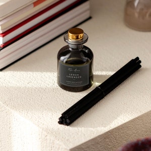 Könnte beinhalten: Eine dunkle Glasflasche mit goldfarbenem Deckel, beschriftet mit "Lemon Lavender Luxury Reed Diffuser". Schwarze Stäbchen sind neben der Flasche angeordnet. Die Flasche steht auf einer weißen Oberfläche.