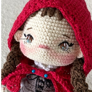May include: Crocheted doll with brown hair, wearing a red hooded cape and a brown and white dress. The doll has blue and brown eyes and a pink blush on its cheeks. The doll is holding a small blue and white bag. @lian2807